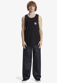 Zwart katoenen tanktop met een borstzakje voorzien van een witte logo. Gecombineerd met brede, donkerblauwe spijkerbroeken en zwarte sneakers.
