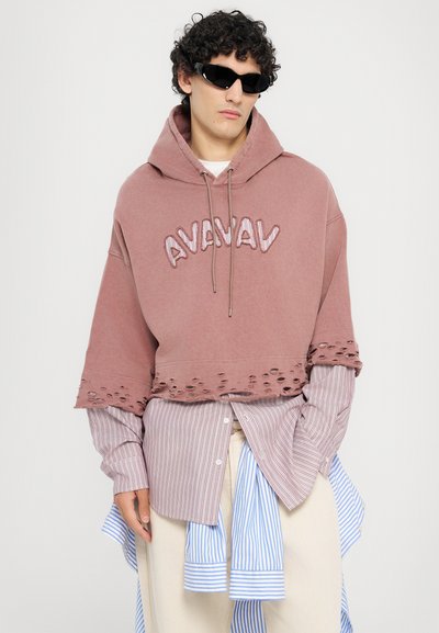 Pink cropped hoodie with distressed hem and embroidered "AVAVAV" text. Layered over a white and blue striped shirt. Black sunglasses.