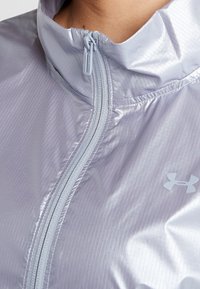Hellblau-lila Sportjacke mit hohem Kragen, Frontreißverschluss, strukturiertem Stoff und kleinem Logo auf der Brust. Hat ein glattes Finish.