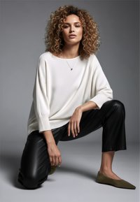 Weißer übergroßer Pullover mit herabgesetzten Schultern, schwarze Fauxlederhosen und olivgrüne Ballerinas, die glatte Texturen und schlichtes Design zeigen.