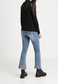 Kobieta ubrana w czarny sweter z koronkowym tyłem, jasnoniebieskie jeansy z postrzępionym dołem oraz czarne sznurowane buty, stojąca tyłem do kamery.