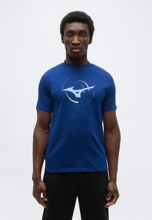 Mann trägt ein königsblaues T-Shirt mit kurzen Ärmeln, bedruckt mit einem großen abstrakten weißen und gelben Logo auf der Brust, und schwarze Hosen, steht vor einem weißen Hintergrund.