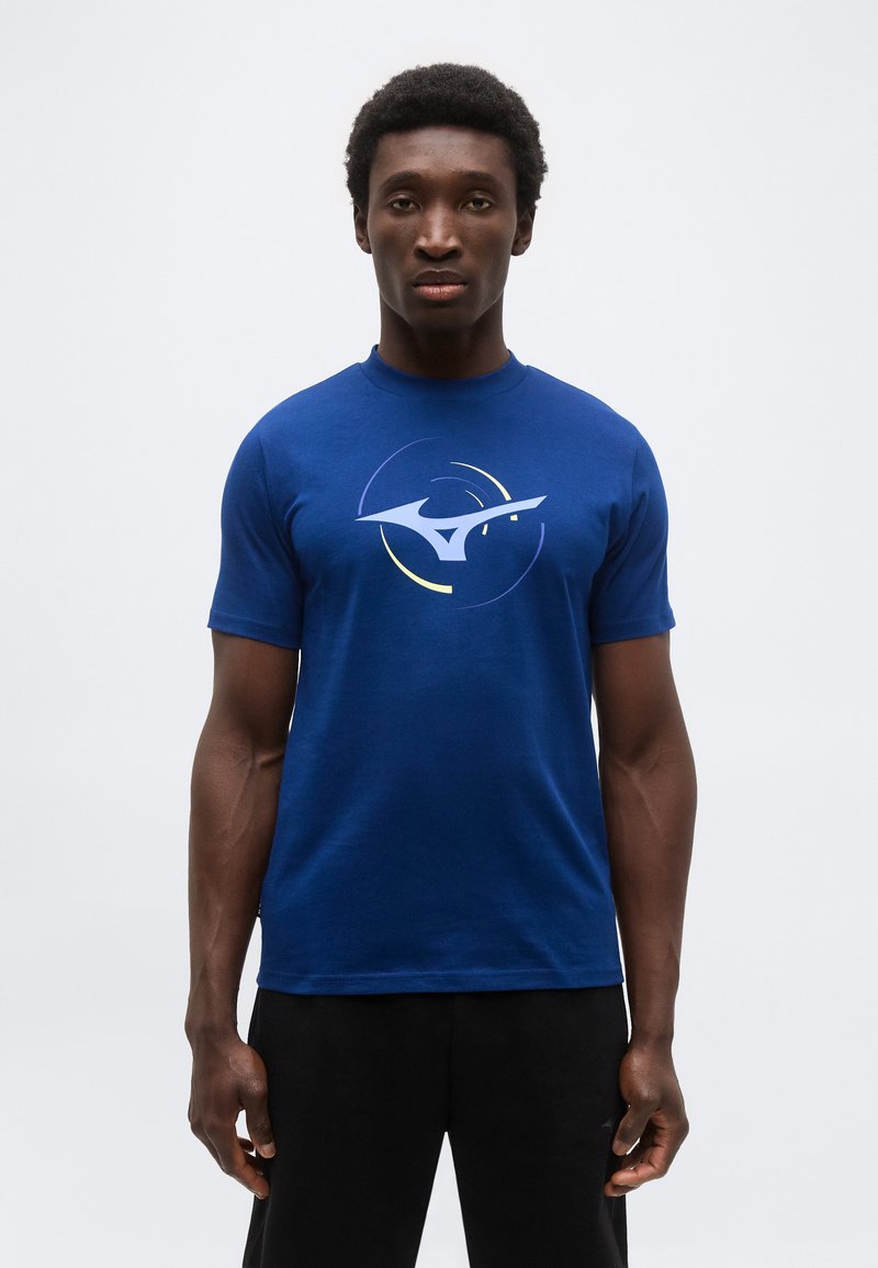 Άνδρας που φοράει βασιλικό μπλε κοντομάνικο μπλουζάκι με μεγάλο αφηρημένο λευκό και κίτρινο λογότυπο στο στήθος και μαύρο παντελόνι, στέκεται μπροστά σε λευκό φόντο.