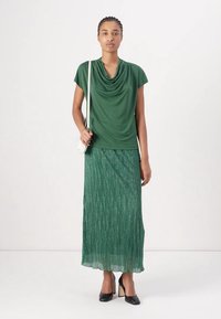 Top verde drappeggiato con maniche corte, abbinato a una gonna maxi verde plissettata e tacchi neri. Include una piccola borsa a tracolla bianca.