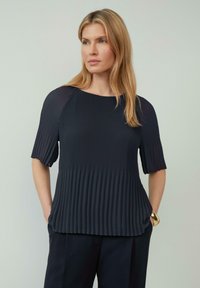 Blouse bleu marine foncé avec une texture plissée, des manches courtes et un col rond, assortie à un pantalon sombre ajusté. Les accents incluent un bracelet en or.