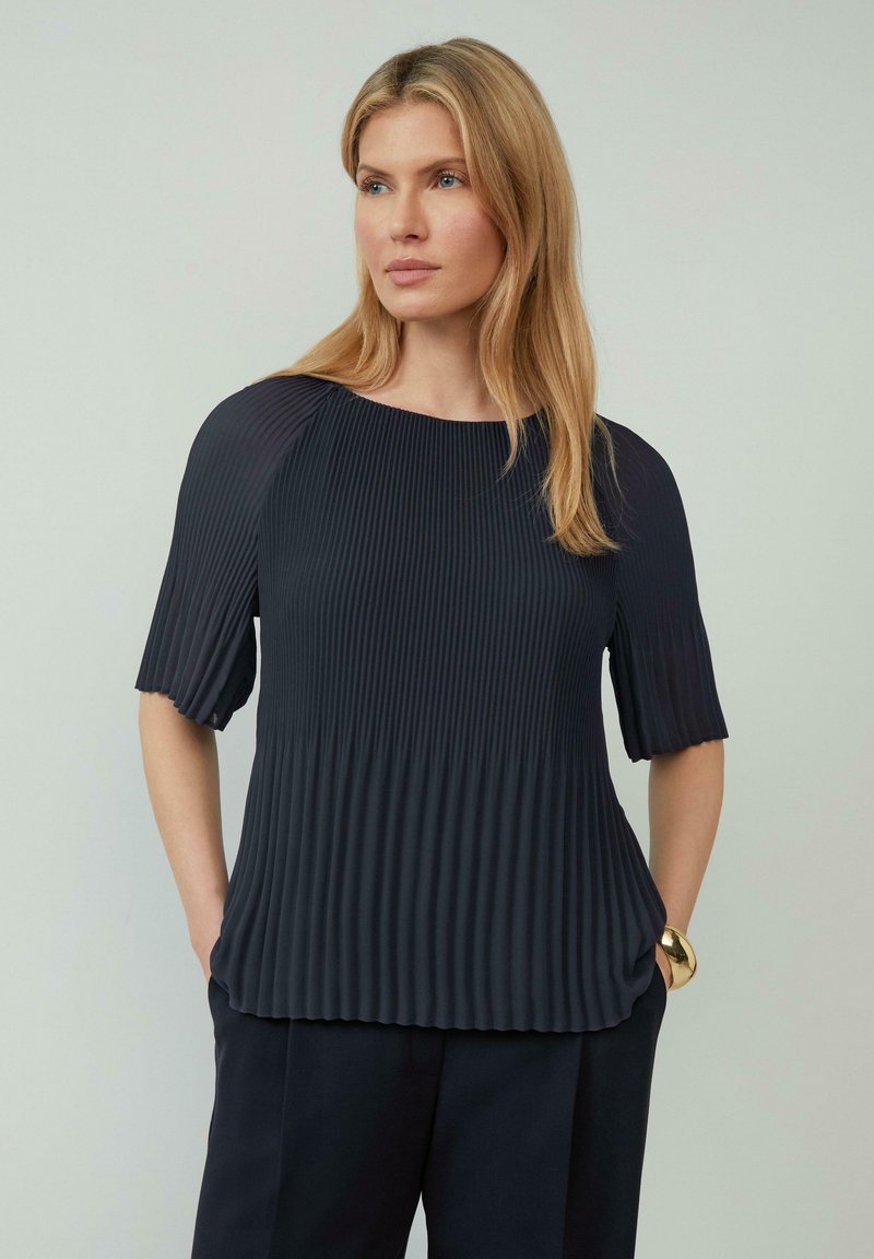 Blouse bleu marine foncé avec une texture plissée, des manches courtes et un col rond, assortie à un pantalon sombre ajusté. Les accents incluent un bracelet en or.