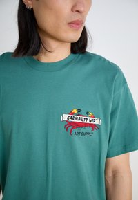 Tyrkysové bavlněné tričko s grafikou kraba a textem "CARHARTT WIP ART SUPPLY" v červené, černé a žluté barvě. Krátké rukávy, kulatý výstřih.