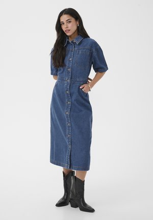 Kvinde med langt mørkt hår iført en knappet denim midi-kjole med korte ærmer og sorte læder cowboystøvler, stående med hænderne i lommerne.