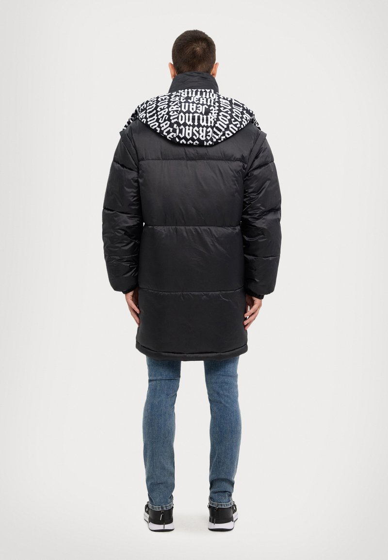 Abrigo de plumas negro con capucha estampada, que presenta un diseño acolchado y mangas largas. Combinado con jeans azules y zapatillas negras.