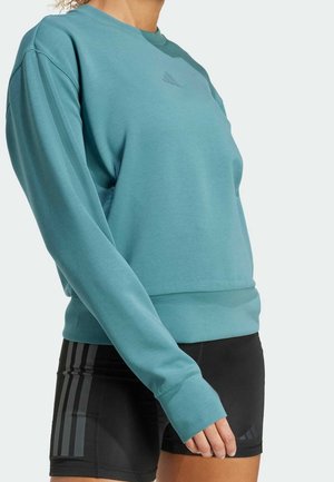 Osoba w turkusowej bluzce z długim rękawem i czarnych sportowych szortach z trzema pionowymi paskami po bokach, widoczna od ramion do połowy uda.