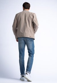 Lichtbeige suède jasje met geribbelde schouderaccenten, gecombineerd met vervaagde blauwe jeans en witte sneakers, gezien vanuit de achterkant.