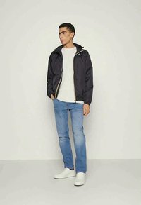 Casaco azul-marinho com fecho de zip e capuz, combinado com uma camisa leve e jeans azuis. O modelo usa ténis brancos. Textura suave.