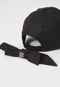 Black baseball cap with a satin tie accent, featuring a logo label. The fabric is smooth with a structured shape and elastic back.