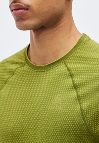 Κοντινό πλάνο ενός ατόμου που φοράει πράσινο μπλουζάκι με υφή και στρογγυλή λαιμόκοψη, με λεπτό λογότυπο στο στήθος.