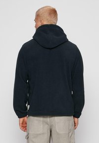 Mann med kort blondt hår iført hettegenser i svart og lysebeige cargo-bukser, sett fra ryggen mot en enkel grå bakgrunn.