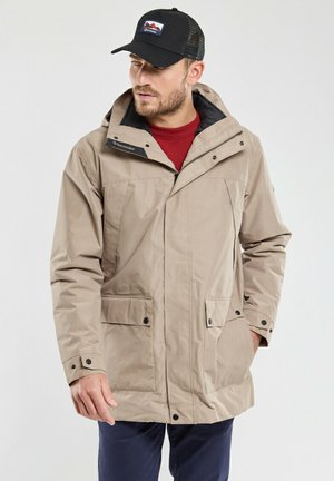 Homme portant une veste à capuche beige sur une chemise rouge, un pantalon bleu marine et une casquette noire avec un logo de montagne, debout devant un fond uni.