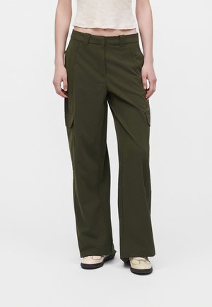 Personne portant un pantalon cargo large vert foncé avec poches à rabat et baskets claires, se tenant devant un fond uni.