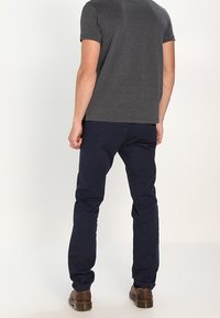 Mann trägt ein dunkelgraues kurzärmliges T-Shirt, eine marineblaue Hose und braune Lederschuhe und steht mit dem Rücken zur Kamera vor einem einfarbigen Hintergrund.