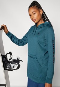 Tyrkysová mikina s kapucí, s přední kapsou, černým logem na prsou a nápisem "BURTON" vytištěným na rukávu, v kombinaci se snowboardem.