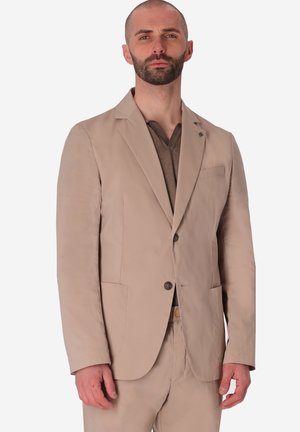 Uomo con barba corta che indossa una giacca beige sopra una maglietta marrone a scollo a V, in piedi con espressione neutra su uno sfondo semplice.