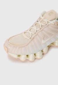 Nike Shox sneaker featuring a light beige mesh upper, curved sole with cushioning columns, and patterned accents on the sides.