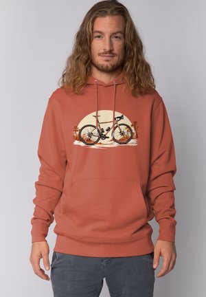 Man met lang haar draagt een roestoranje hoodie met een afbeelding van een racefiets, palmbomen en een zonsondergang op de achtergrond.
