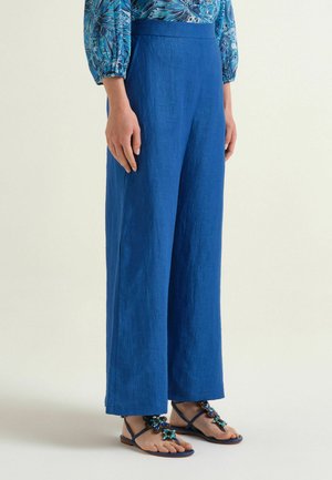 Femme portant un pantalon large bleu vif, un chemisier bleu fleuri à manches bouffantes et des sandales bleu marine avec des motifs floraux ornés de bijoux.