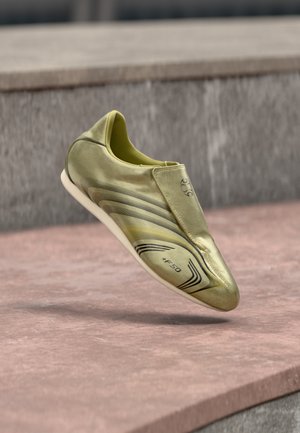 Arany focicipő, elegáns dizájnnal, sima szintetikus anyagból, bebújós kialakításban, fekete díszítő vonalakkal, amelyek a "+F50" logót tartalmazzák.