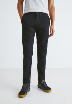 Uomo che indossa pantaloni casual neri e sneakers nere con suola gialla, in piedi con le mani parzialmente nelle tasche su uno sfondo bianco uniforme.