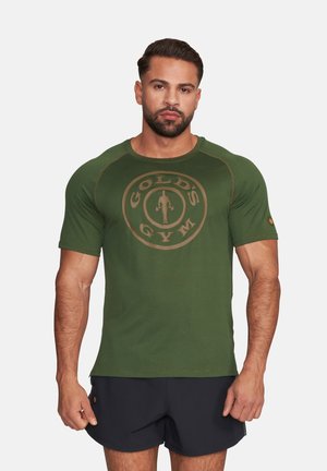 Muskelbepackter Mann mit kurzen, dunklen Haaren und Bart trägt ein grünes Gold's Gym Shirt und schwarze Shorts, steht vor einfarbigem Hintergrund.