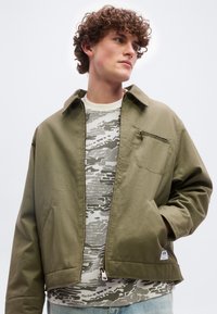 Νέος άνδρας με σγουρά μαλλιά, φορώντας ένα χακί τζάκετ με φερμουάρ πάνω από ένα λευκό και γκρι πουκάμισο με σχέδια, κοιτάζει προς τα πλάγια με το ένα χέρι στην τσέπη.