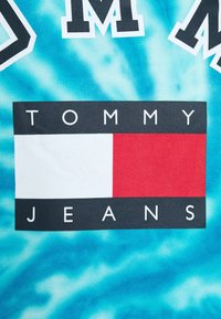 Blå tie-dye tyg har en rektangulär logotyp med "TOMMY" ovanför och "JEANS" nedanför, satt på en svart bakgrund med en vit och röd ruta.