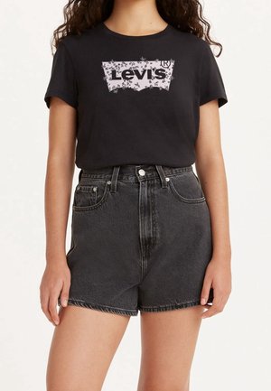 Kvinde iført sort Levi's t-shirt med blomsterlogo og højtaljede, falmede sorte denimbukser, stående mod en ensfarvet lys baggrund.
