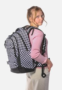 Černo-bílý károvaný batoh s více přihrádkami a černými akcenty, nošený přes růžový svetr s dlouhými rukávy.