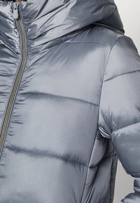 Giacca imbottita grigia con finitura lucida, dotata di chiusura con cerniera, cappuccio e pannelli cuciti per l'isolamento termico. Tessuto liscio con design aderente.