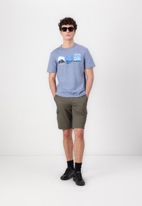 Lys blå bomull T-skjorte med fotografiske trykk; olivengrønne cargosshorts; svarte joggesko; mørke solbriller. Minimale tilbehør, uformell passform.