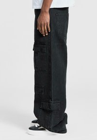 Persona che indossa pantaloni cargo neri a gamba larga e sneaker bianco e neri, in posizione laterale contro uno sfondo chiaro e semplice.