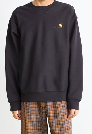 Personne portant un sweat-shirt Carhartt noir avec un petit logo sur la poitrine et un pantalon à carreaux marron, debout devant un fond blanc.