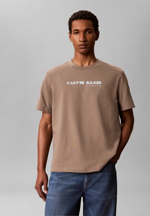 Młody mężczyzna w jasno-brązowej koszulce Calvin Klein i niebieskich dżinsach, stojący z jedną ręką w kieszeni na szarym tle.