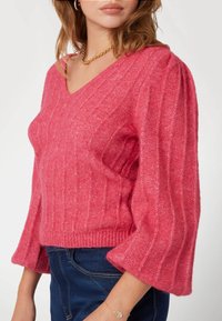 Maglione rosa lavorato a maglia con scollatura a V, texture a coste e maniche a campana. Il tessuto appare morbido ed elastico, creando una silhouette rilassata.