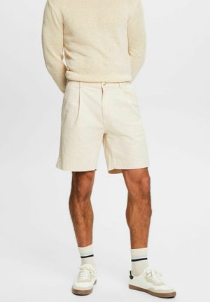 Homme portant un pull en tricot beige, un short plissé blanc cassé, des chaussettes blanches avec des rayures noires et des baskets blanches, debout sur un fond blanc.