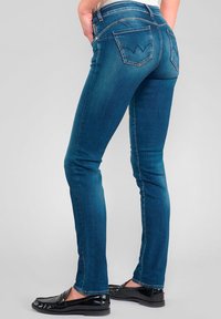 Jean skinny en denim bleu avec une taille mi-haute, deux poches arrière présentant un design en chevron, et ajusté de la taille à la cheville.