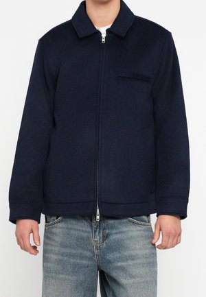 Osoba ubrana w granatową kurtkę zapinaną na zamek błyskawiczny z kołnierzem i kieszenią na piersi, w połączeniu z jasnoniebieskimi dżinsami.