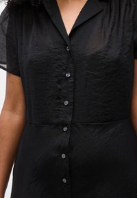 Κοντινό πλάνο ενός ατόμου που φοράει μαύρο φόρεμα με κοντά μανίκια και υφή, με κουμπιά και γιακά σε σχήμα V.