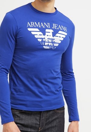 Mies pukeutuneena istuvaan siniseen pitkähihnaiseen paitaan, jossa valkoinen "Armani Jeans" -teksti ja kotkalogo, yhdistettynä tummiin farkkuihin.