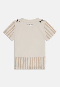 Beige kortärmad skjorta med ljusbruna vertikala ränder på ärmar och fåll, svarta Puma-logotyper på axlarna och texten "KidSuper" nära kragen.