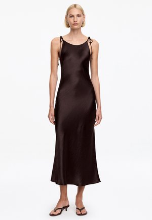 Kobieta ubrana w ciemnobrązową, bezrękawną sukienkę do kostek z cienkimi, wiązanymi ramiączkami i czarne sandały na obcasie.