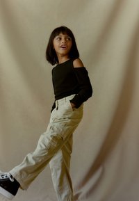 Zwarte off-shoulder top met lange mouwen, gecombineerd met beige cargobroek met zakken. Het model draagt zwarte sneakers en poseert tegen een neutrale achtergrond.