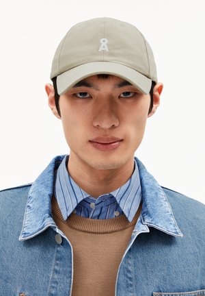 Khaki baseball cap with white embroidered logo on front. Worn with a denim jacket, blue striped shirt, and brown sweater.