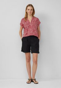 Blouse à motifs rouges avec des manches courtes, associée à un short noir roulé et des sandales plates sur un fond neutre.
